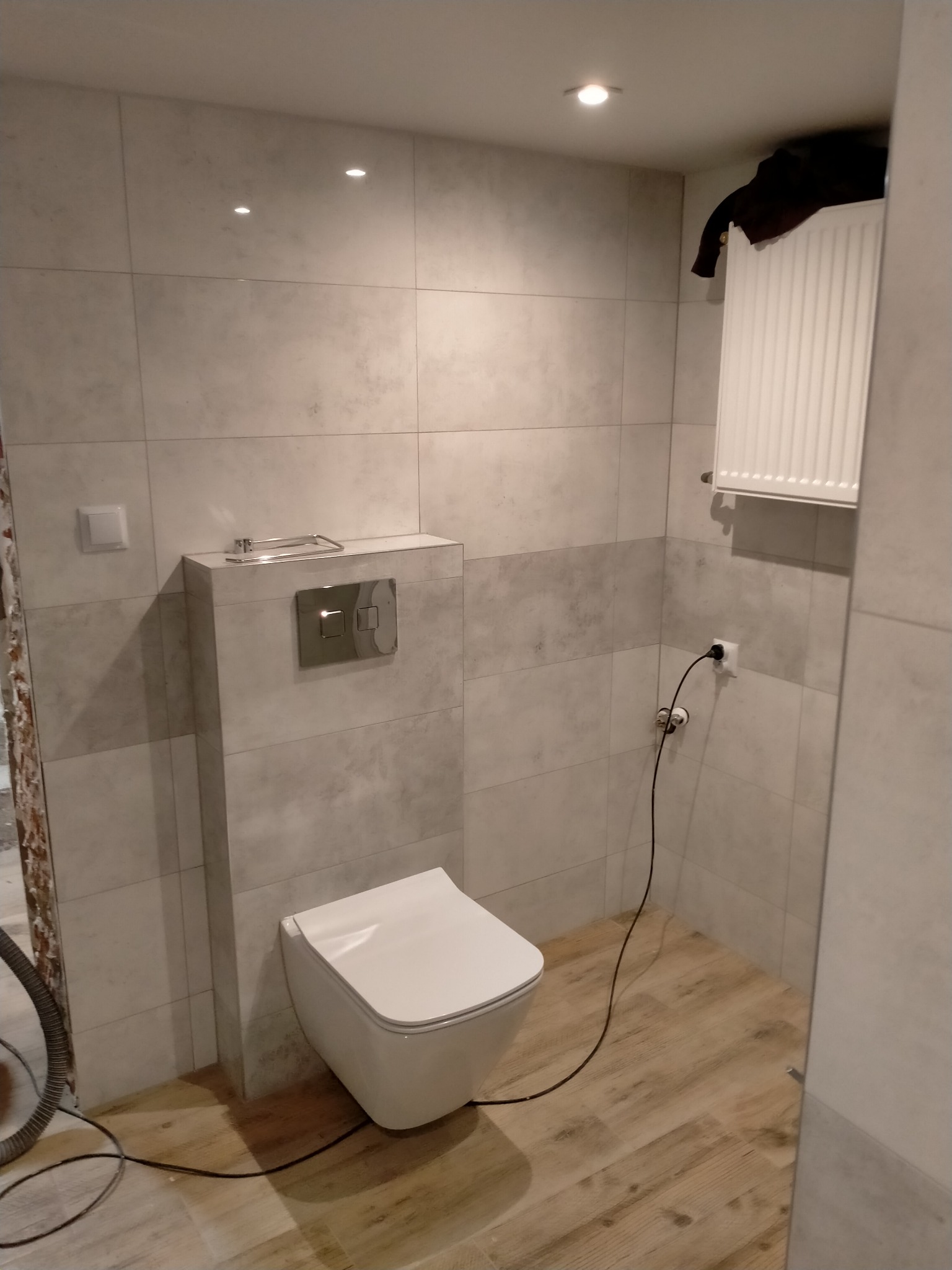 Nowoczesna toaleta w trakcie montażu w łazience wykończonej płytkami imitującymi kamień, widoczny fragment odkurzacza przemysłowego i przewody elektryczne.