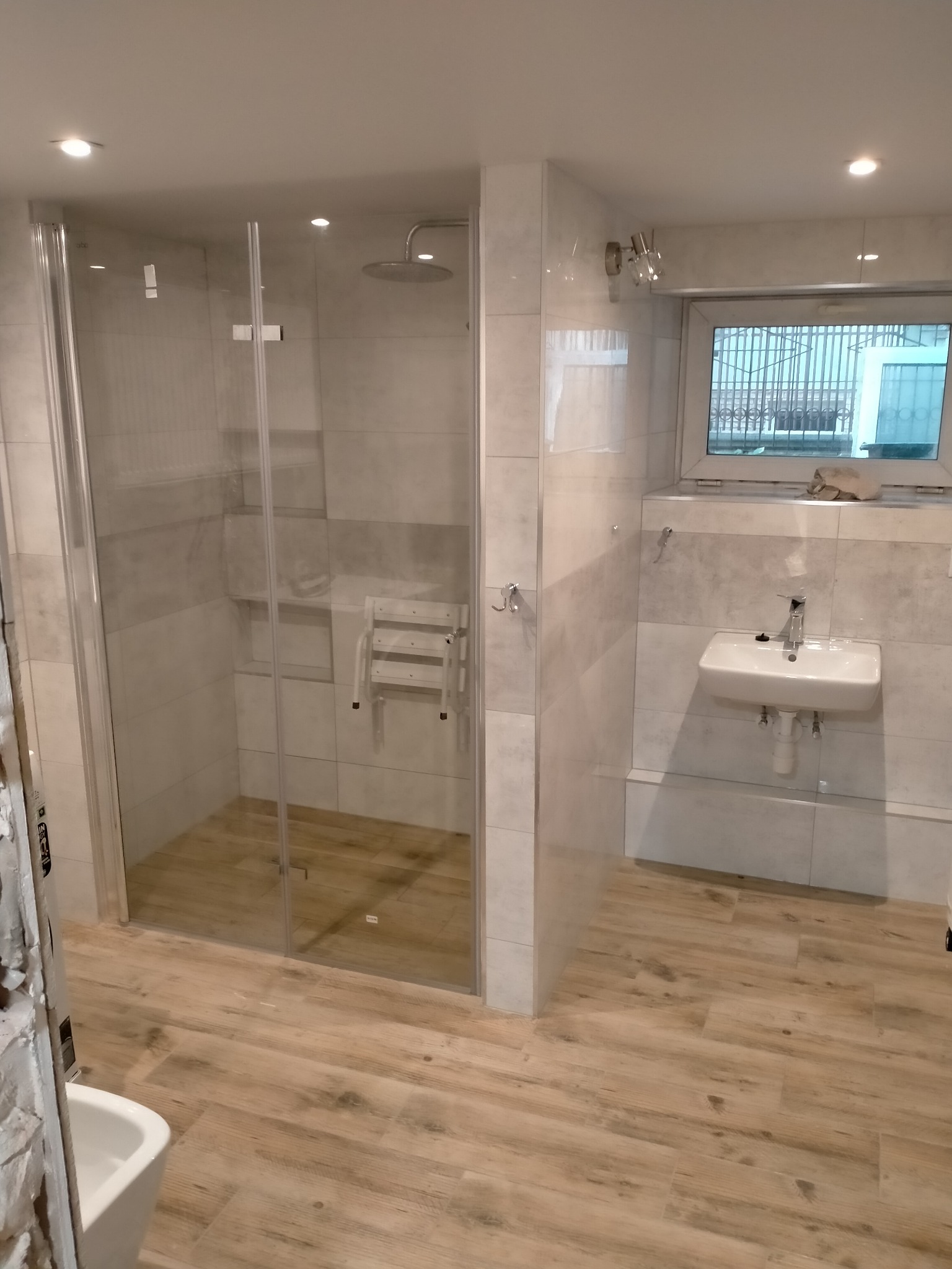 Nowoczesna łazienka z kabiną prysznicową typu walk-in, wykończona jasnymi płytkami imitującymi drewno na podłodze i szarymi płytkami na ścianach, z zamontowanym składanym siedziskiem wewnątrz...