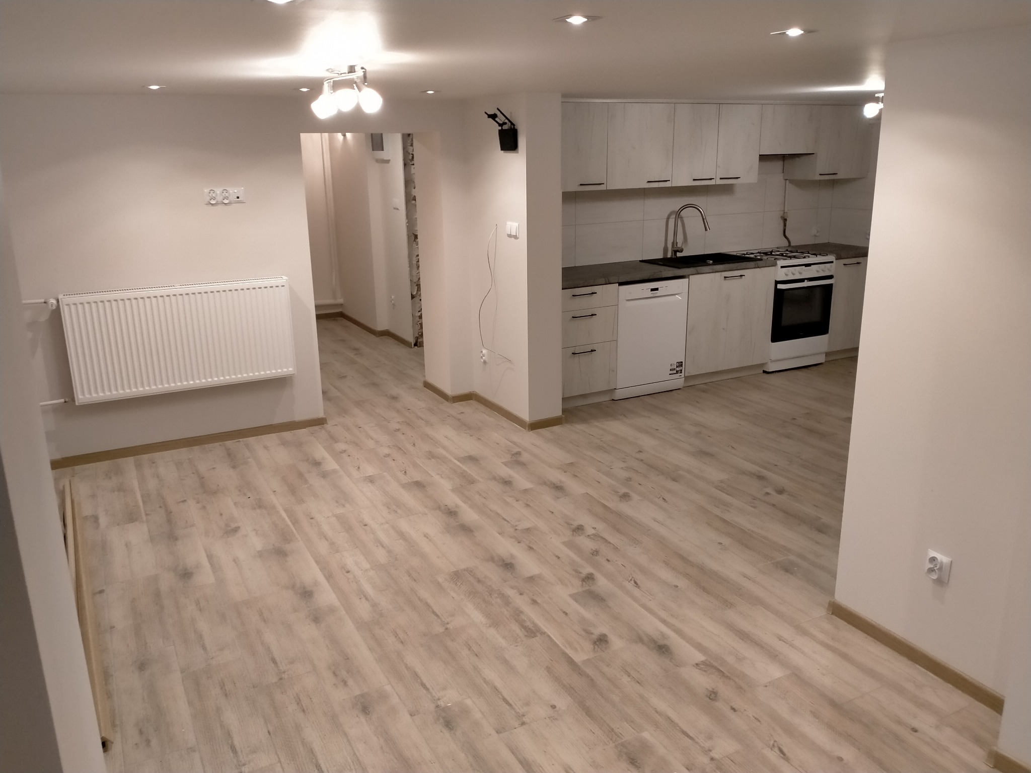 Wykończone wnętrze mieszkania z panelami podłogowymi, białymi ścianami, grzejnikiem i widoczną kuchnią w tle, oświetlone halogenami.