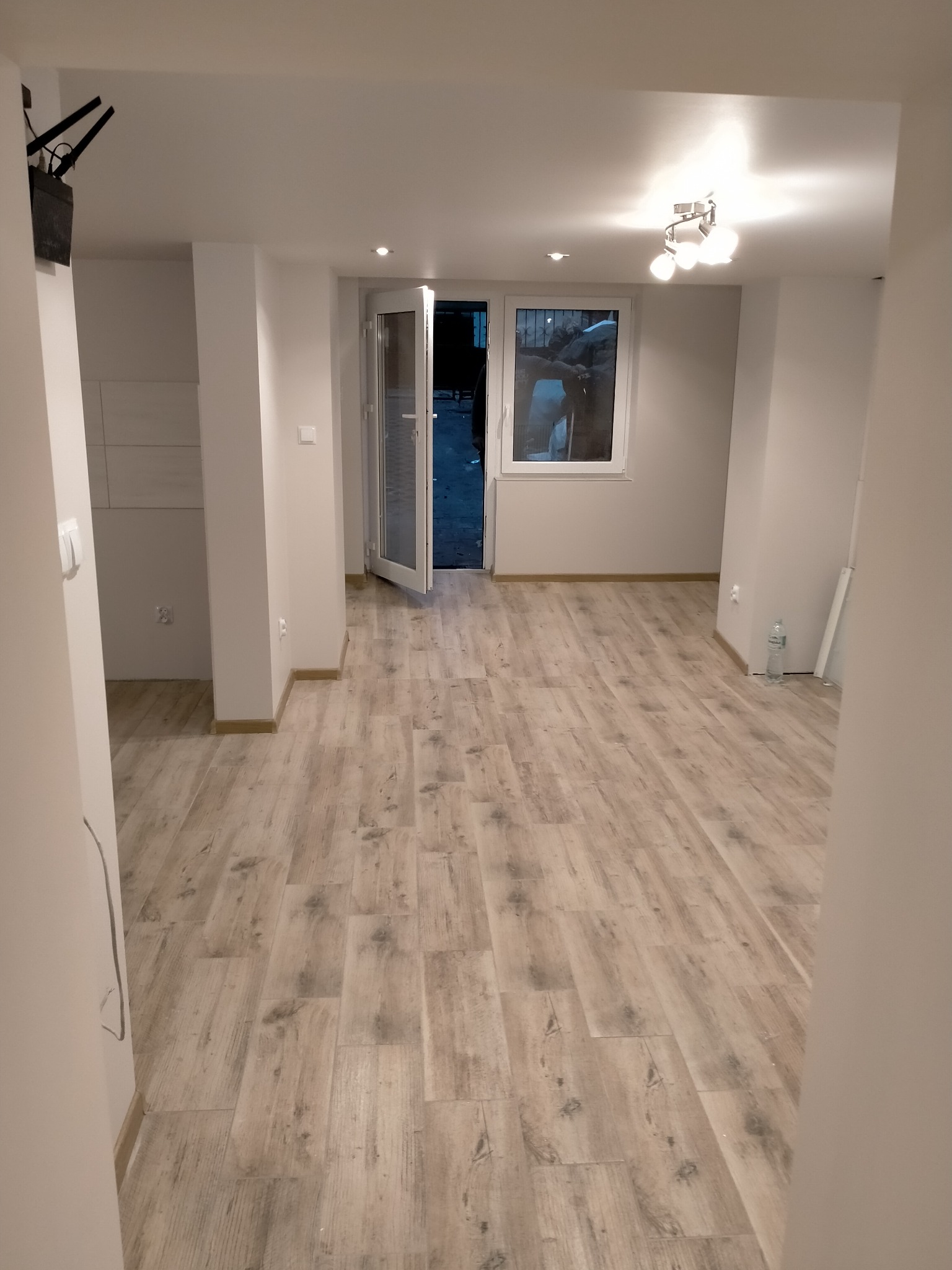 Puste, odświeżone wnętrze mieszkania z drewnopodobną podłogą, białymi ścianami i otwartymi drzwiami balkonowymi, widok z korytarza.