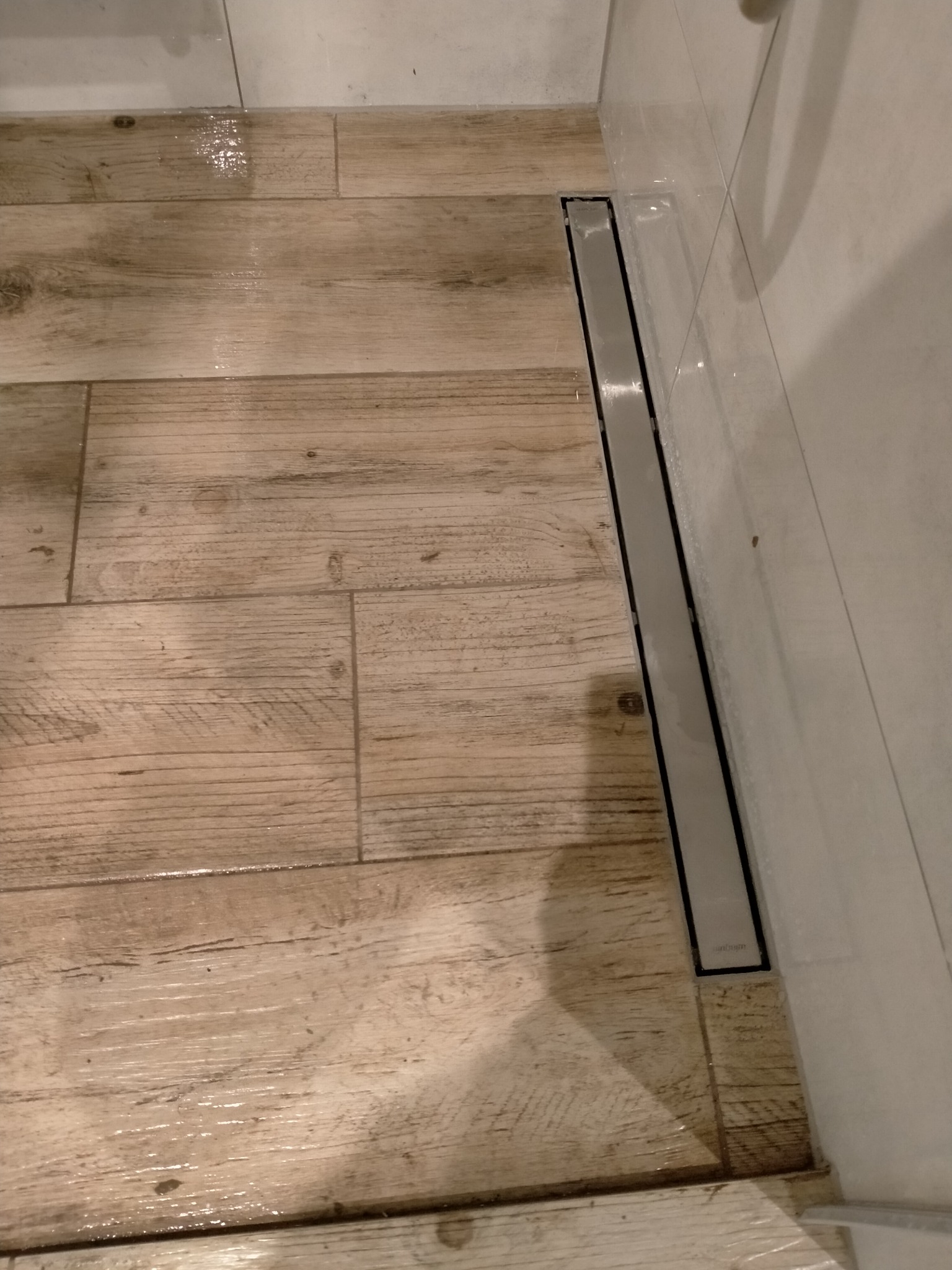 Podłoga w kabinie prysznicowej wyłożona płytkami imitującymi drewno, z odpływem liniowym przy ścianie, lekko mokra po użyciu.