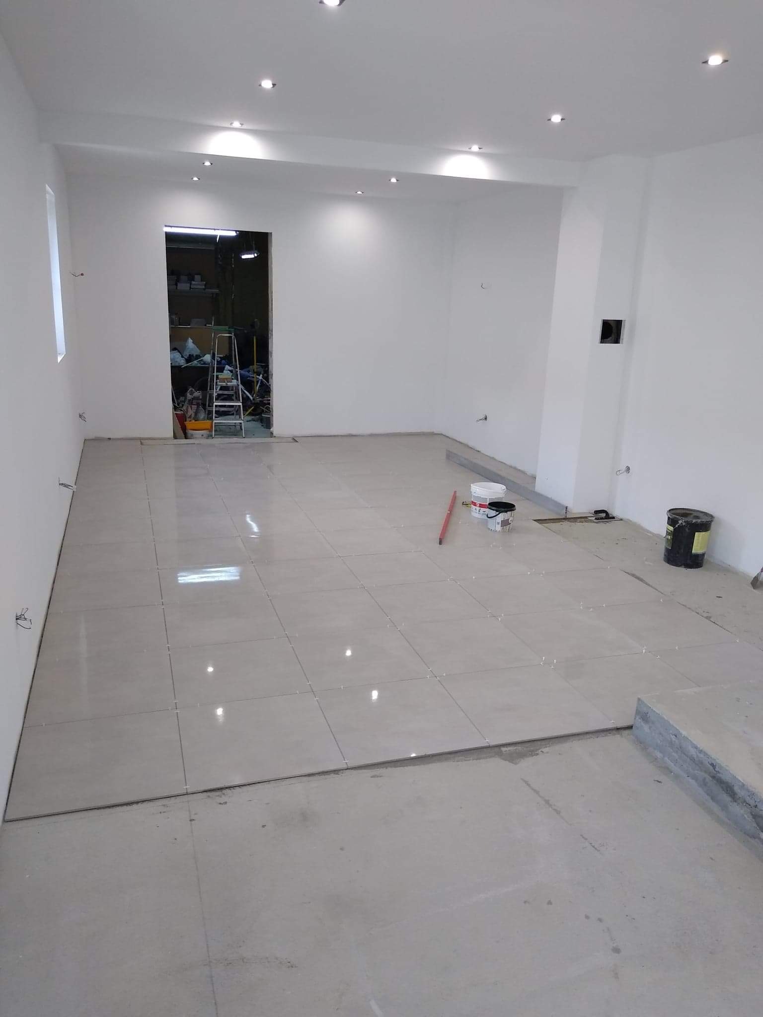 Wnętrze w trakcie układania jasnych płytek podłogowych; widoczne narzędzia, poziomica, wiaderka z zaprawą, krzyżyki dystansowe i fragment nieukończonej powierzchni.