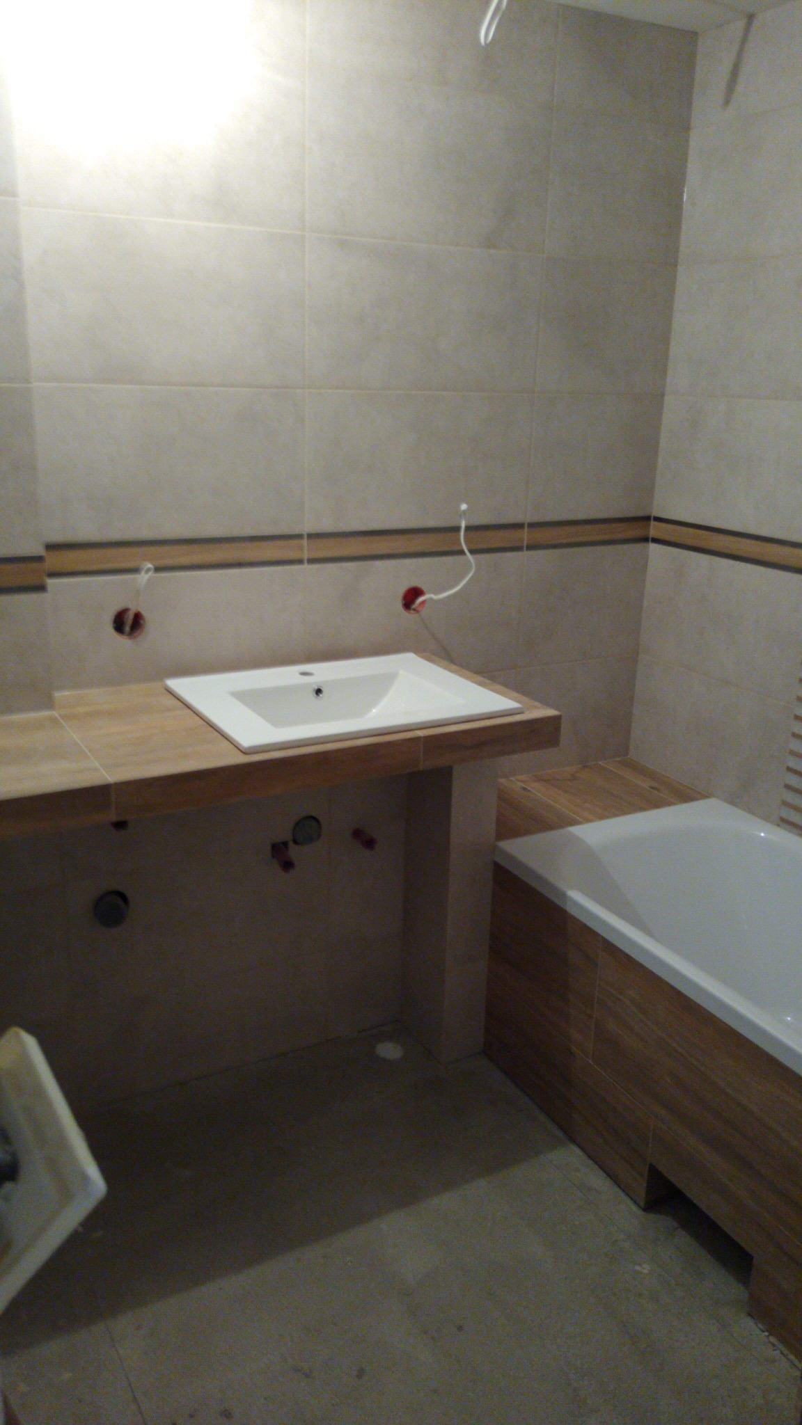 Łazienka w trakcie prac wykończeniowych: umywalka nablatowa na drewnianym blacie, obudowana wanna, płytki na ścianach, widoczne instalacje hydrauliczne i elektryczne przed montażem armatury.