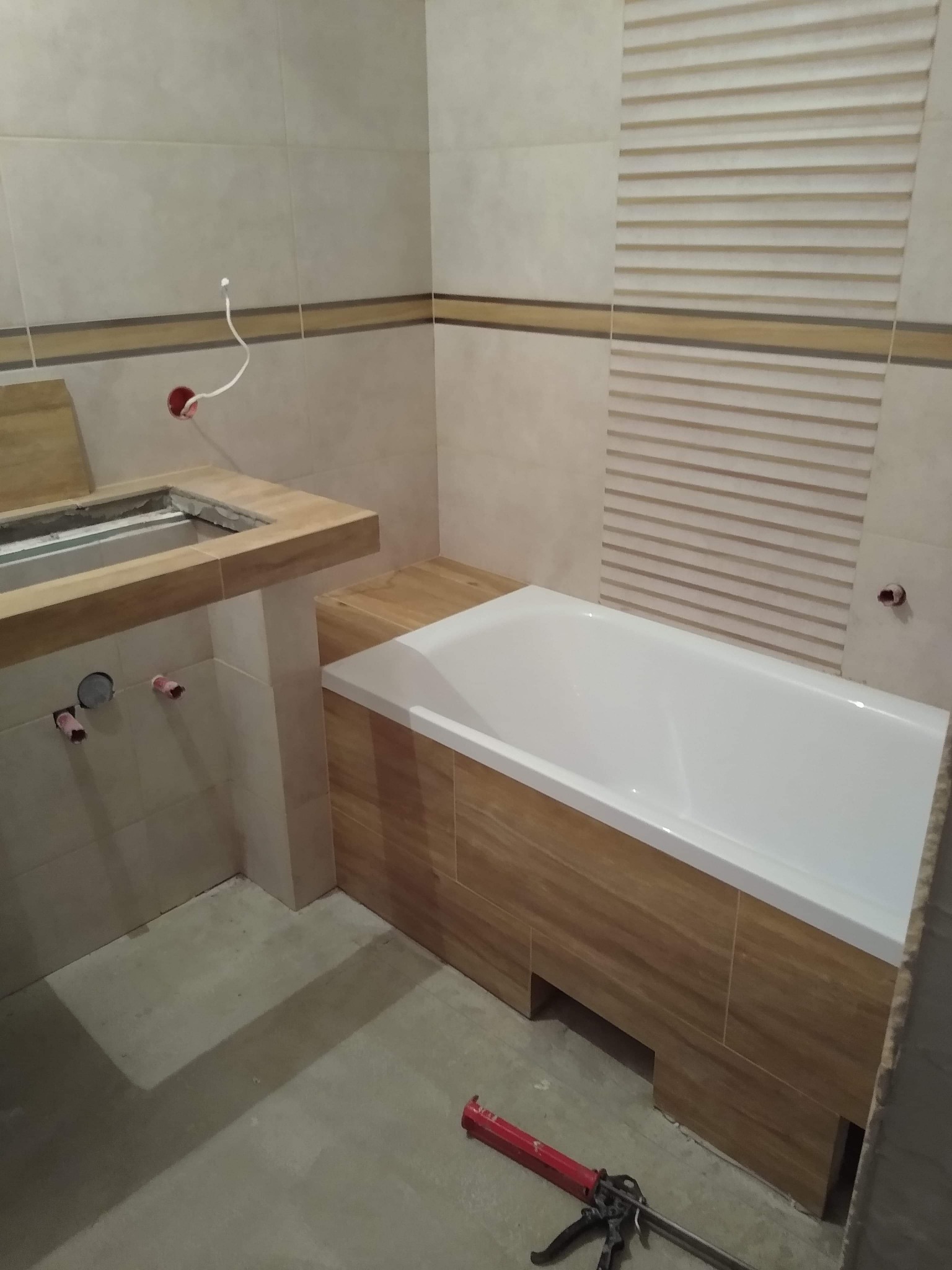 Niedokończona łazienka z białą wanną obudowaną panelami imitującymi drewno, umywalką z blatem w trakcie montażu, beżowymi płytkami na ścianach i pistoletem do silikonu na podłodze.