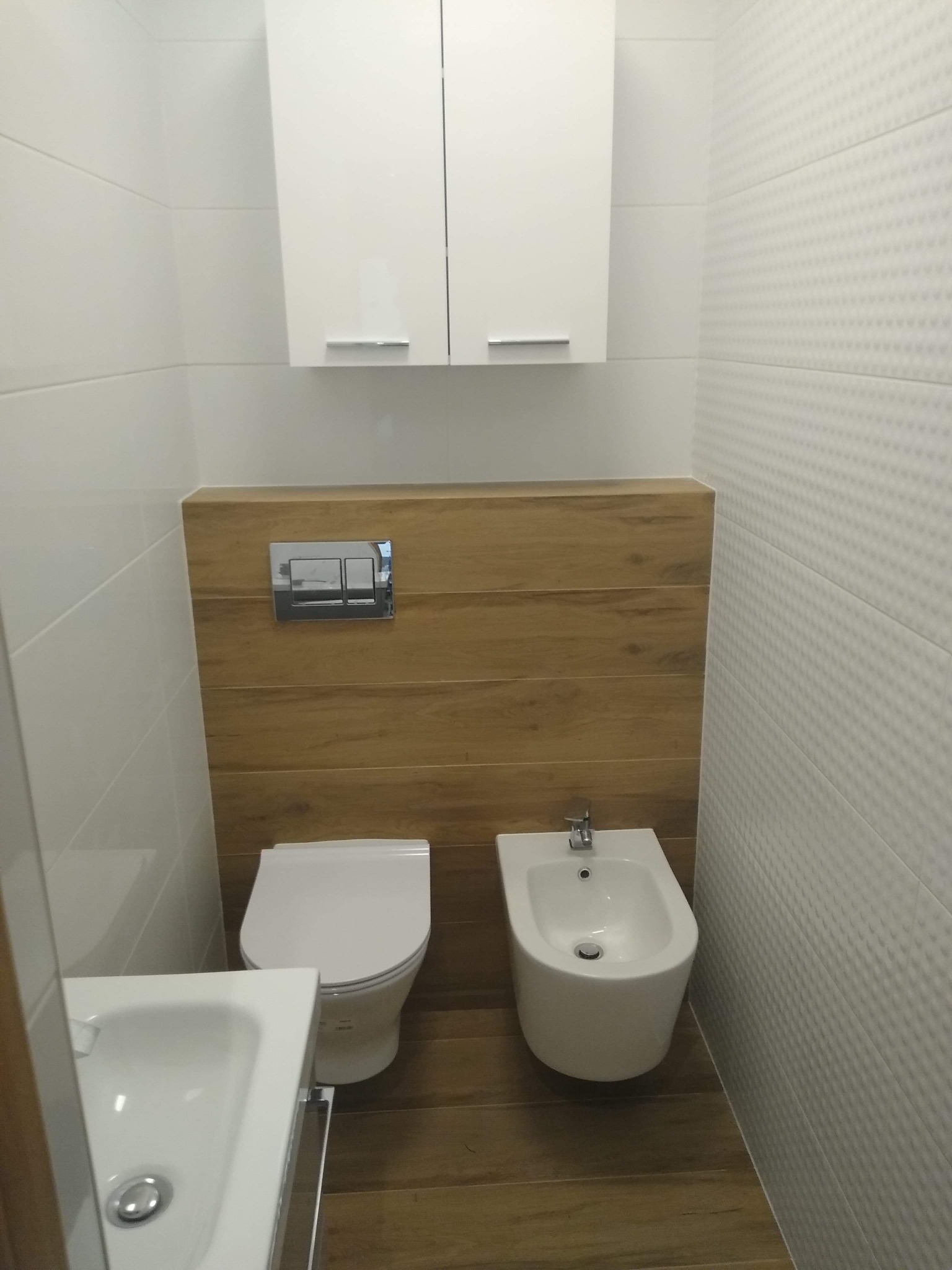 Nowoczesna łazienka z białymi płytkami o fakturze rombów, drewnopodobną zabudową, wiszącą szafką i białymi sanitariatami: toaleta i bidet.