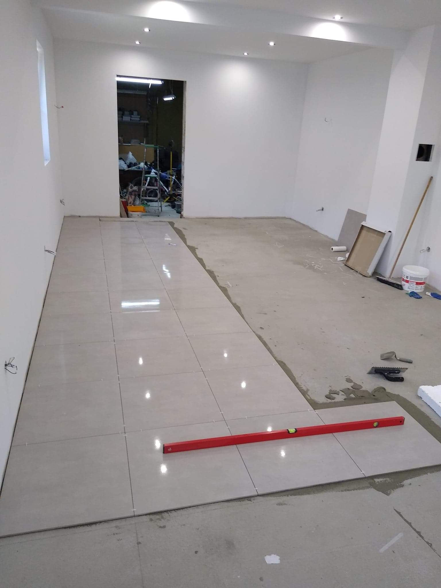 Układanie jasnych, kwadratowych płytek podłogowych w trakcie prac wykończeniowych, widoczny poziomica i narzędzia glazurnicze, surowy beton na nieukończonej części podłogi.