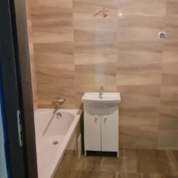 Wykończona łazienka z beżowymi płytkami imitującymi kamień na ścianach i podłodze, biała wanna i szafka pod umywalką, widoczne narzędzia glazurnicze na ścianie.