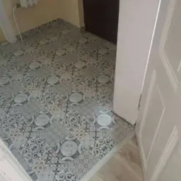 Wykończona podłoga w pomieszczeniu z wzorzystymi płytkami w odcieniach szarości i beżu, widoczne fragmenty drzwi i ściany.