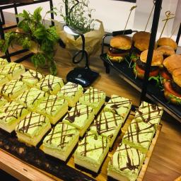 Craft Food Project - Aranżacja cateringu: rzędy kwadratowych ciast z limonkowym kremem i czekoladową polewą obok mini burgerów z wykałaczkami, oświetlone lampką biurkową, uzupełnione roślinami doniczkowymi.
