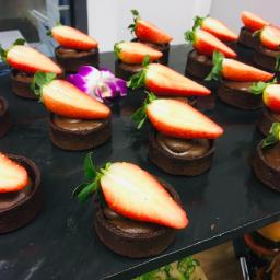 Craft Food Project - Rząd czekoladowych tartaletek z kremem i połówkami truskawek na czarnym talerzu, z dekoracyjnym fioletowym kwiatem.