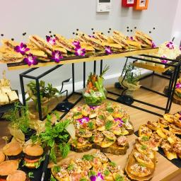 Craft Food Project - Bogato zastawiony stół z różnorodnymi przekąskami: mini burgery, kanapki z dekoracją z fioletowych kwiatów, bruschetty z szynką parmeńską i ziołami, oraz warzywa w szklanym naczyniu.