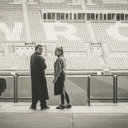 Craft Food Project - Dwie osoby rozmawiają na tle trybun stadionu, widok z poziomu murawy, czarno-biała fotografia.