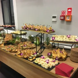 Stół cateringowy z różnorodnymi przekąskami na drewnianych deskach i czarnych łupkach, udekorowany fioletowymi orchideami, z widocznymi kanapkami, wrapami, tartaletkami i deserami, w tle biała...