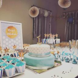 Craft Food Project - Stół z dekoracjami i słodkościami na pierwsze urodziny, w tym dwupoziomowy tort z cyfrą '1', babeczki z niebieskim kremem, ciasteczka i upominki dla gości w przezroczystych woreczkach, w tle...