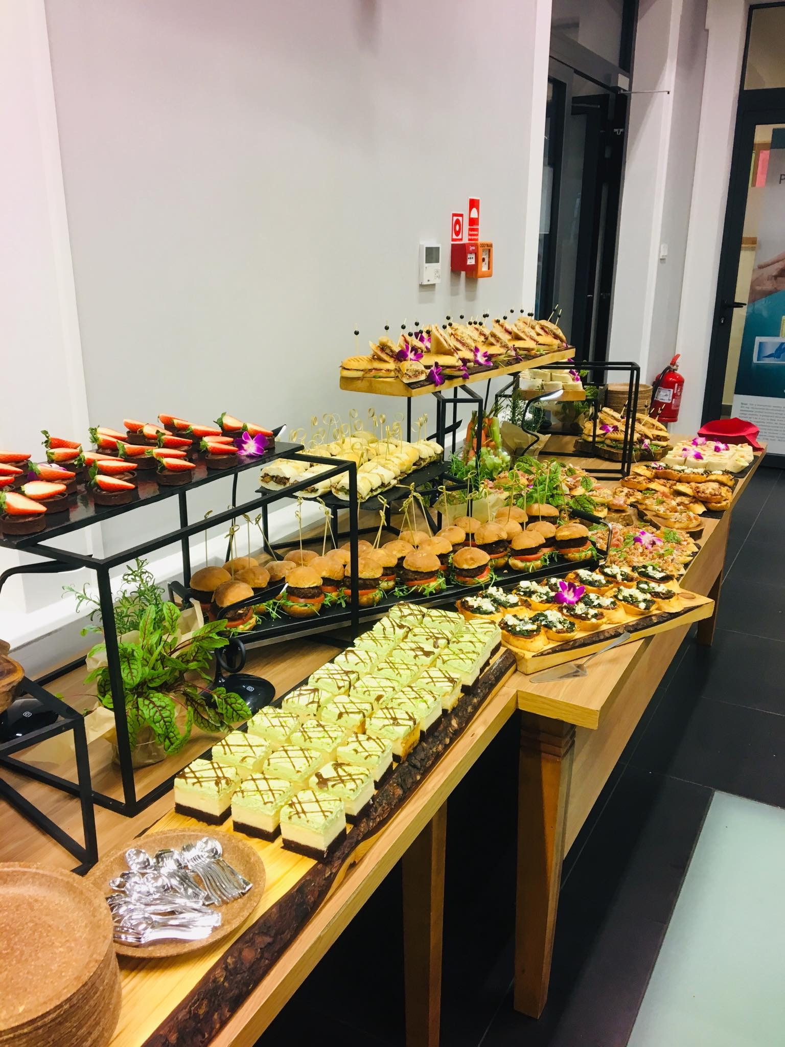 Bogato zastawiony stół z różnorodnymi przekąskami: mini burgery, tartaletki z truskawkami, kanapki, ciasta z zieloną polewą, bruschetta, dekorowane kwiatami, ustawione na drewnianym stole...