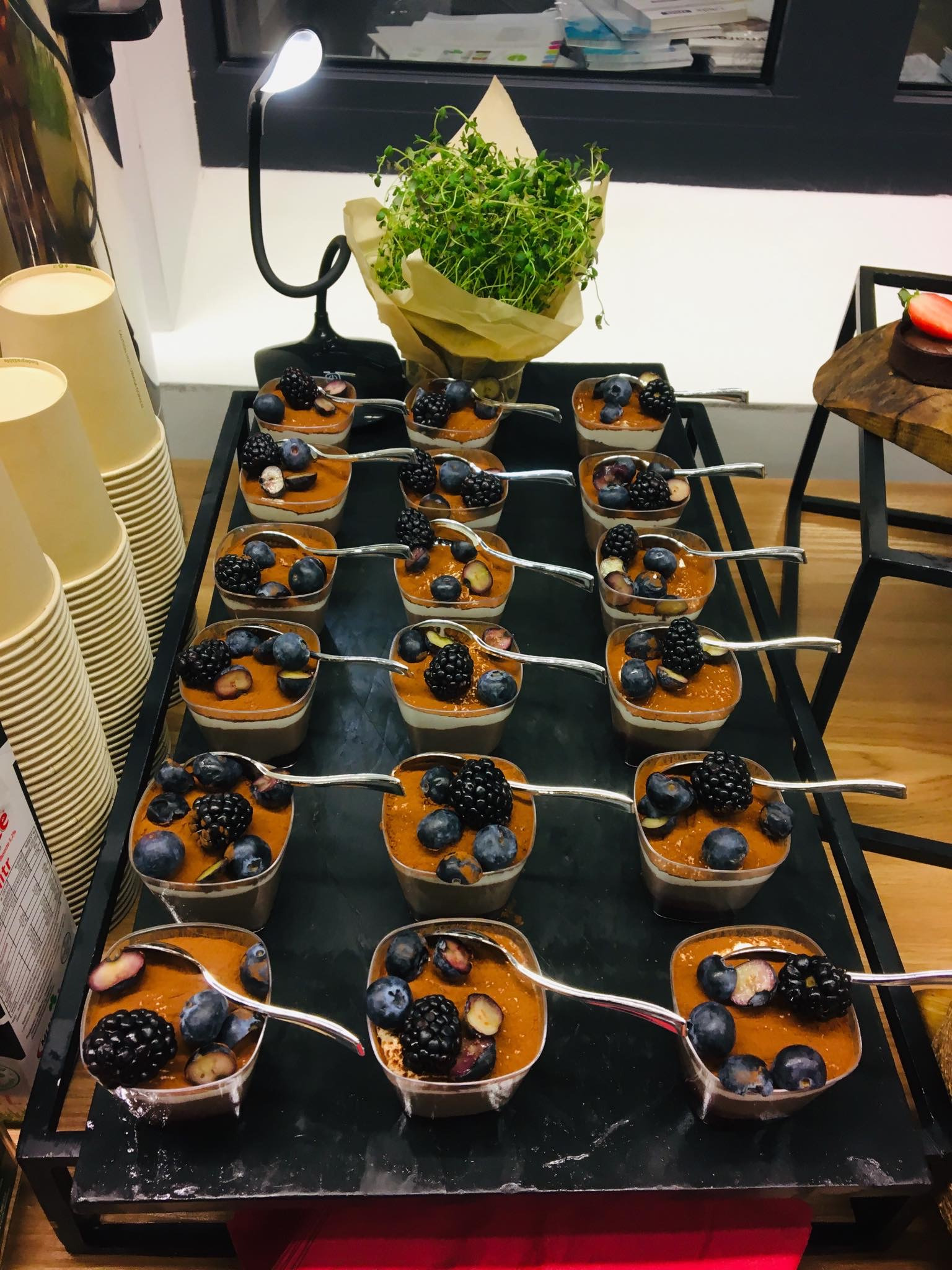 Deserki w małych pojemnikach z kremem, borówkami i jeżynami, ułożone na czarnej tacy obok stosu papierowych kubków i dekoracji z ziół, oświetlone lampką.