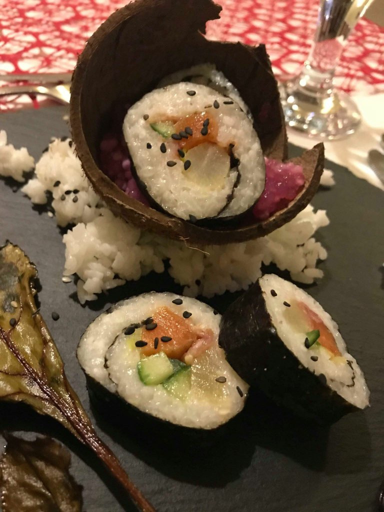 Ujęcie z góry: sushi z łososiem, ogórkiem i czarnym sezamem, podane w skorupie kokosa na czarnym talerzu z ryżem i liściem buraka.