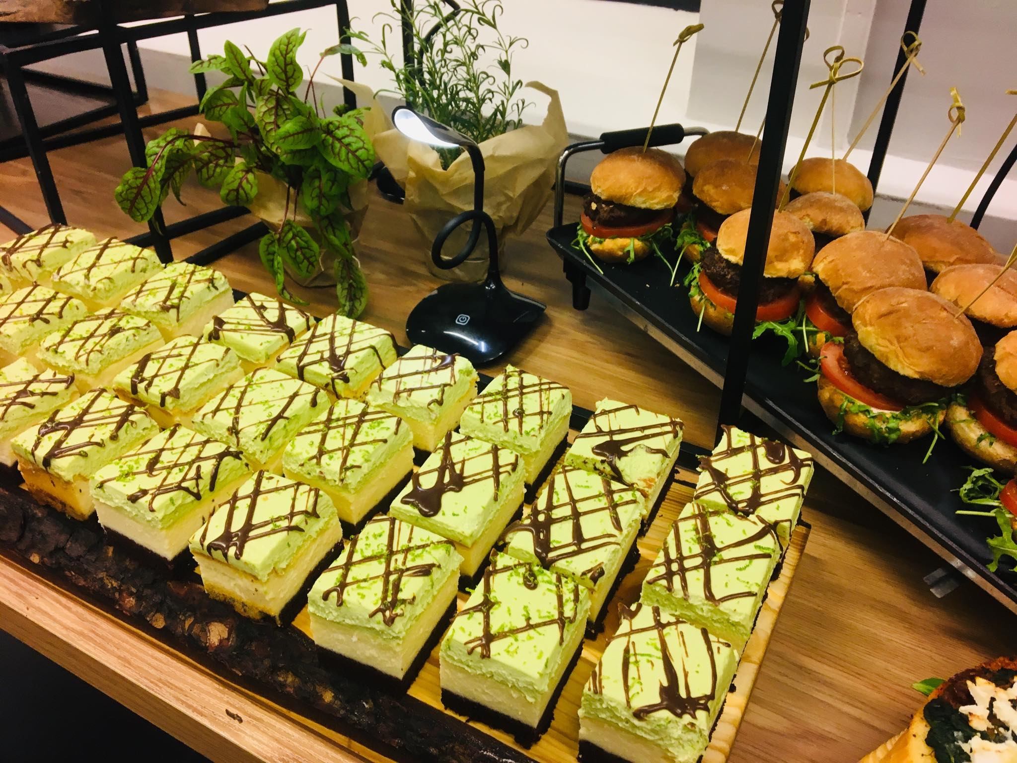 Aranżacja cateringu: rzędy kwadratowych ciast z limonkowym kremem i czekoladową polewą obok mini burgerów z wykałaczkami, oświetlone lampką biurkową, uzupełnione roślinami doniczkowymi.
