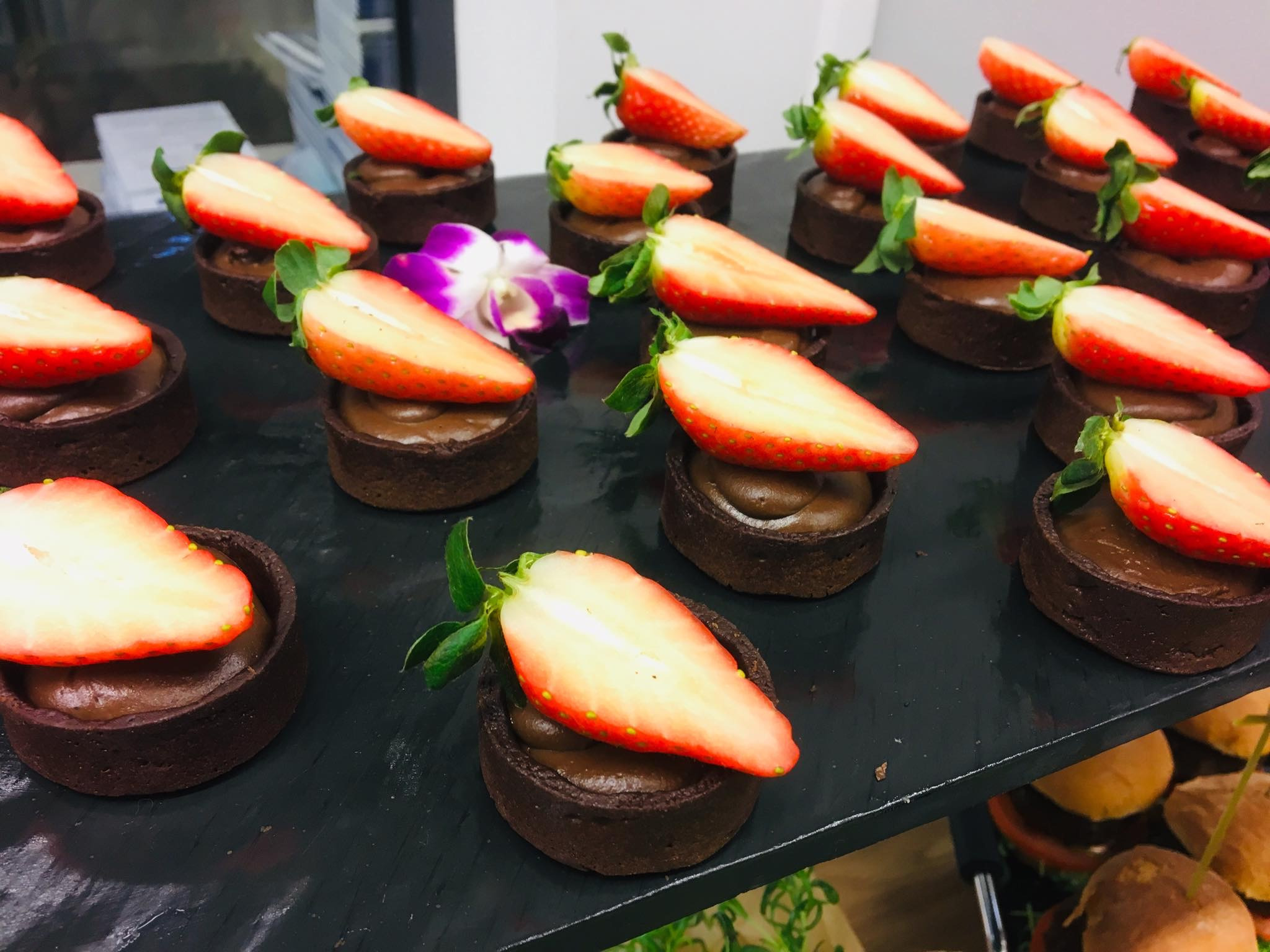 Rząd czekoladowych tartaletek z kremem i połówkami truskawek na czarnym talerzu, z dekoracyjnym fioletowym kwiatem.