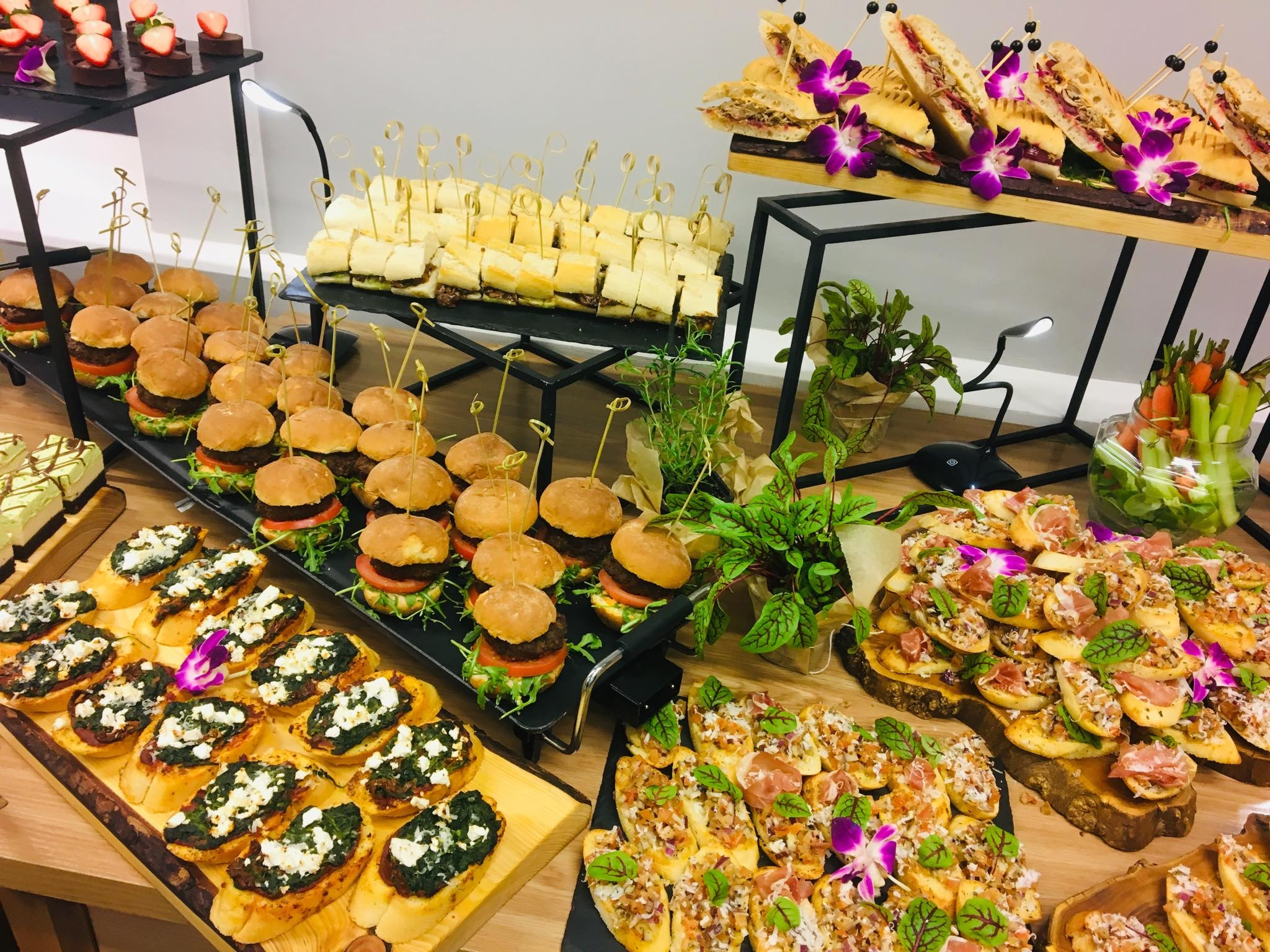 Stół z różnorodnymi przekąskami na przyjęcie: mini burgery, kanapki, bruschetty z pesto i serem, tartaletki z truskawkami, udekorowane świeżymi ziołami i kwiatami, na czarnych i drewnianych tacach.