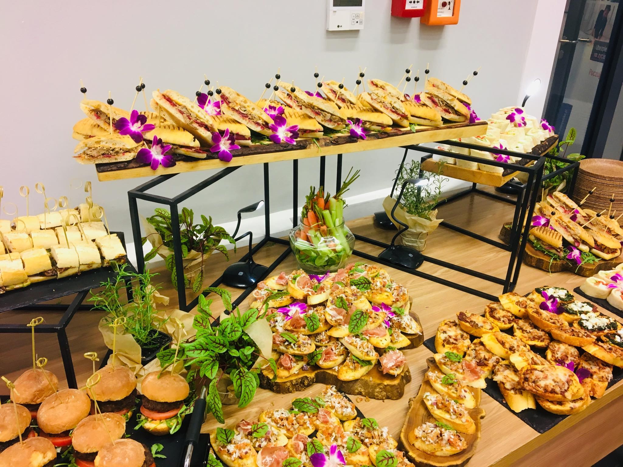 Bogato zastawiony stół z różnorodnymi przekąskami: mini burgery, kanapki z dekoracją z fioletowych kwiatów, bruschetty z szynką parmeńską i ziołami, oraz warzywa w szklanym naczyniu.