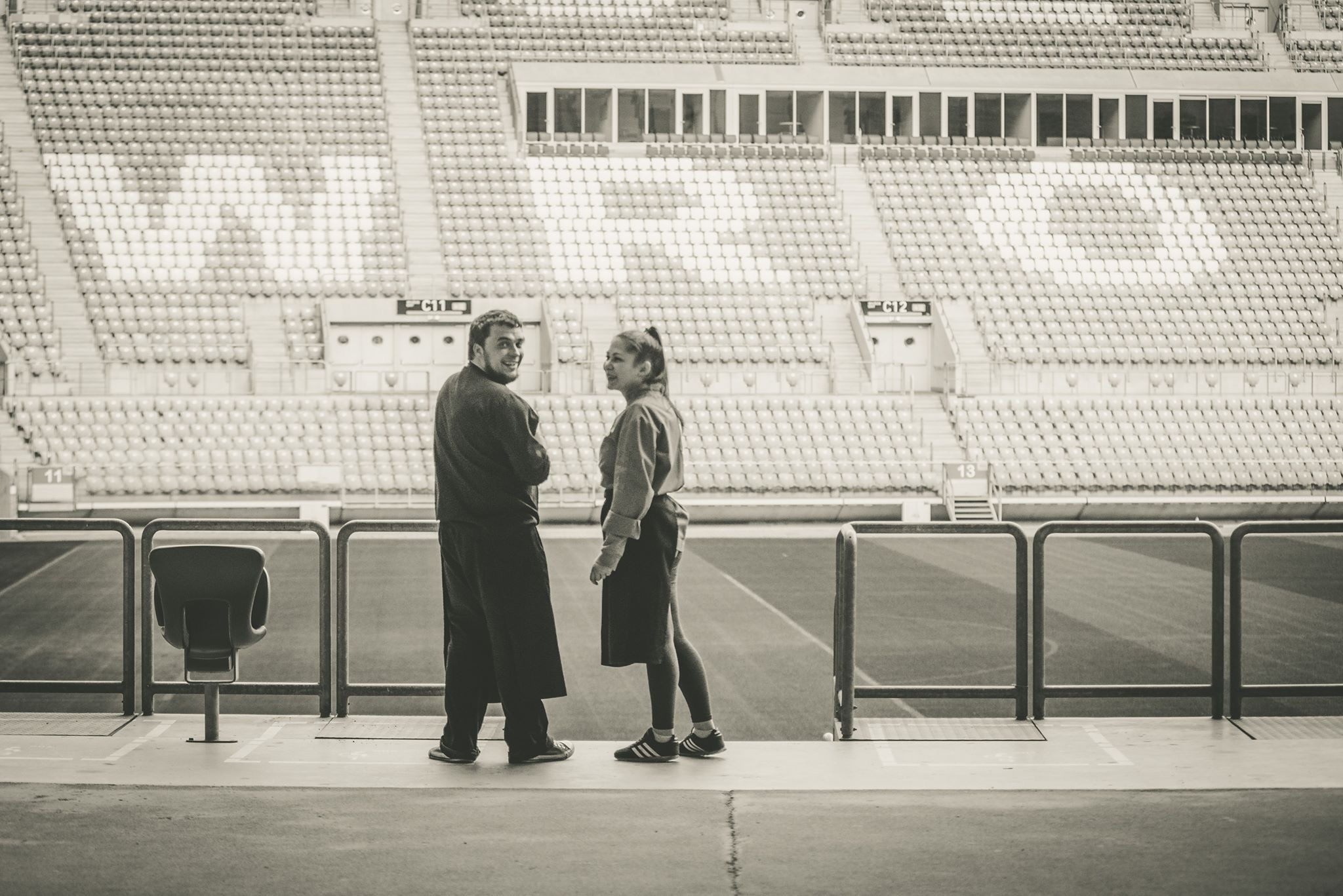 Dwie osoby rozmawiają na tle trybun stadionu, widok z poziomu murawy, czarno-biała fotografia.