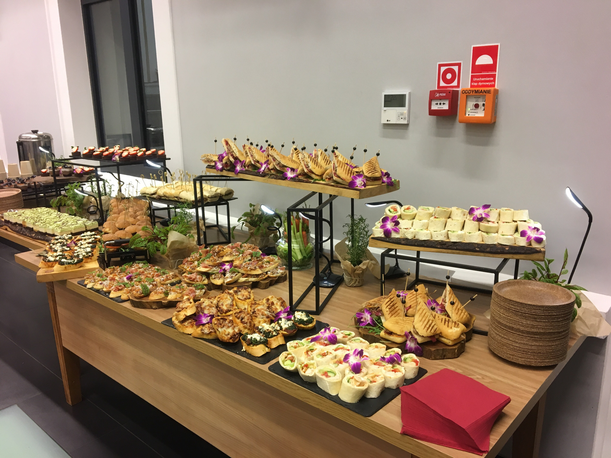 Stół cateringowy z różnorodnymi przekąskami na drewnianych deskach i czarnych łupkach, udekorowany fioletowymi orchideami, z widocznymi kanapkami, wrapami, tartaletkami i deserami, w tle biała...