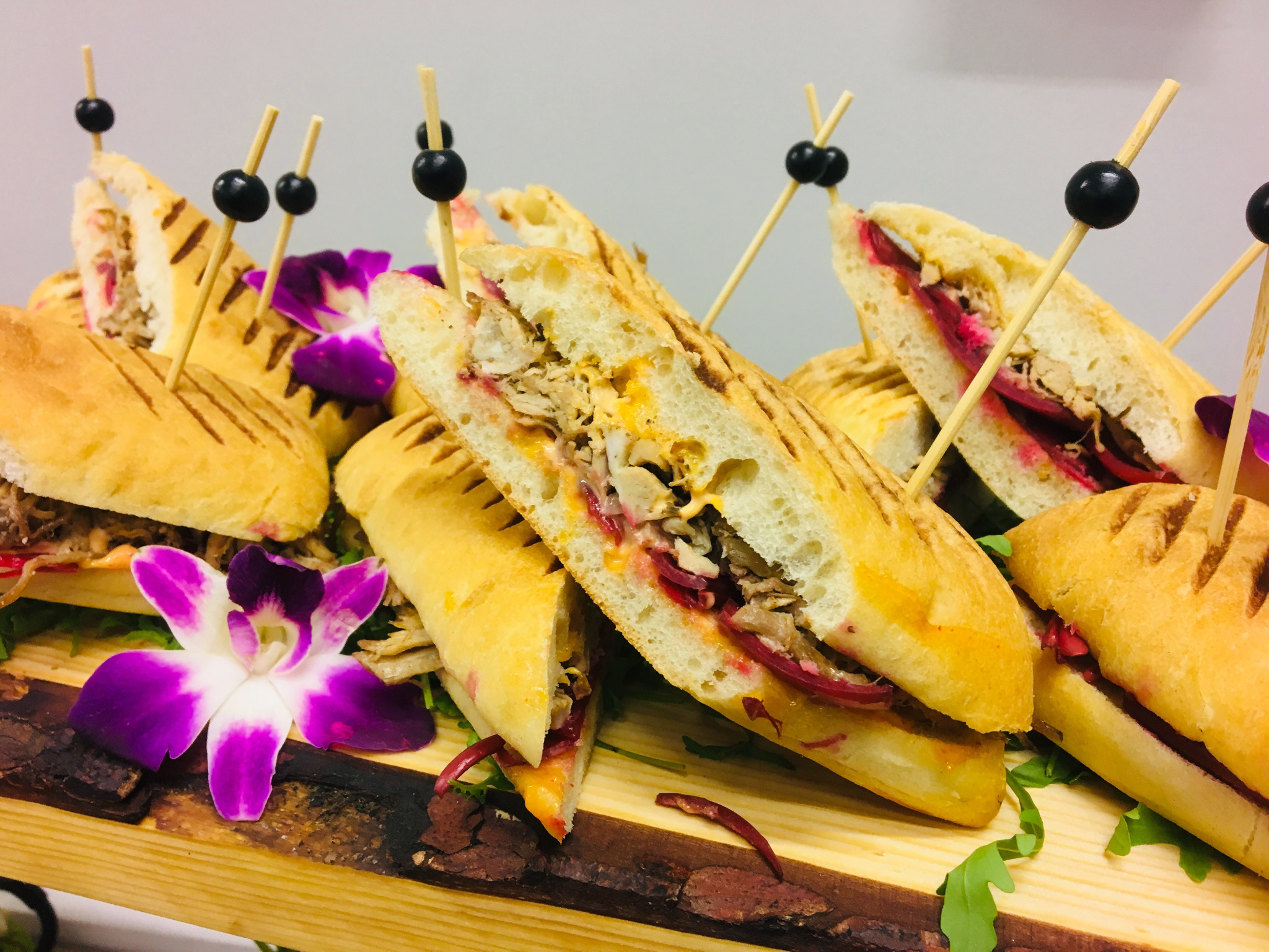 Elegancko podane, przekrojone kanapki z różnymi nadzieniami, ozdobione fioletowymi kwiatami i spięte ozdobnymi wykałaczkami na drewnianej desce.