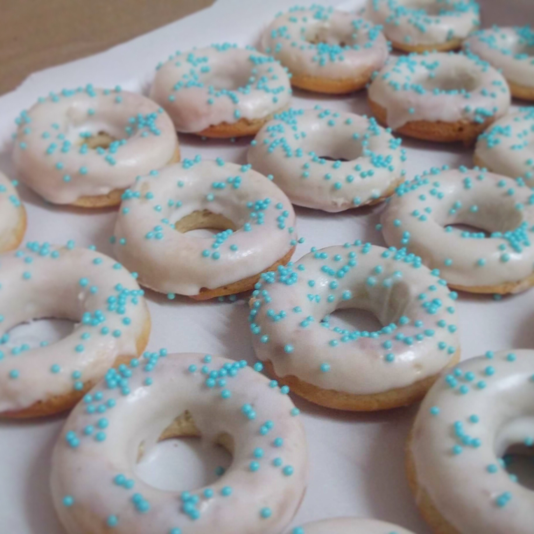 Bliski plan na szereg pączków z białym lukrem i turkusowymi posypkami, ułożonych na białym papierze.