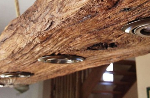 Lampa sufitowa z surowego drewna z trzema punktowymi źródłami światła, widoczne sęki i naturalna faktura drewna, schody w tle.