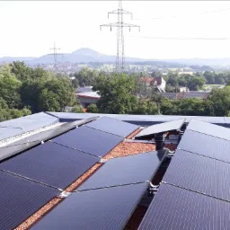 Widok z góry na panele słoneczne na dachu, z widoczną konstrukcją montażową i drobnym żwirem między panelami, w tle krajobraz miejski z liniami wysokiego napięcia.