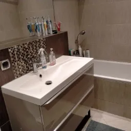 Umywalka z szafką w odcieniach beżu, bateria umywalkowa, płytki ścienne w beżowym kolorze, akcesoria łazienkowe na blacie, wanna w tle.