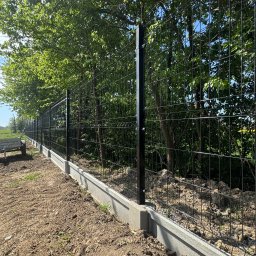 WIKO-MET - Nowoczesne, czarne ogrodzenie panelowe na betonowej podmurówce, widoczne na tle zieleni drzew. Elementy montażowe słupków. Świeżo wyrównana ziemia.