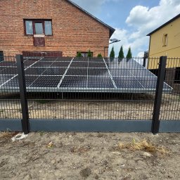 WIKO-MET - Nowoczesne, grafitowe ogrodzenie panelowe z widocznymi panelami słonecznymi w tle, na tle ceglanego domu z oknami. Ujęcie z bliska, skupione na detalu ogrodzenia.