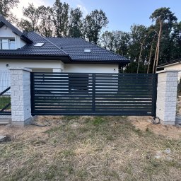 WIKO-MET - Nowoczesna, przesuwna brama wjazdowa z poziomych profili, osadzona na murowanych słupkach z jasnego kamienia. W tle dom jednorodzinny i drzewa.