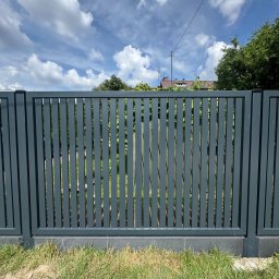 WIKO-MET - Nowoczesne, grafitowe ogrodzenie z pionowych paneli metalowych na niskiej podmurówce, z widokiem na błękitne niebo z chmurami i zieleń w tle.