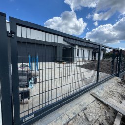 WIKO-MET - Nowoczesna, przesuwna brama wjazdowa z metalową siatką na tle domu z garażem i błękitnego nieba z chmurami. Widoczne elementy montażowe i teren w trakcie prac wykończeniowych.