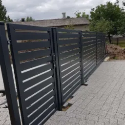 Nowoczesna, przesuwna brama wjazdowa z poziomych profili w kolorze antracytowym, zamontowana na posesji z kostką brukową, widoczny fragment domu w tle.