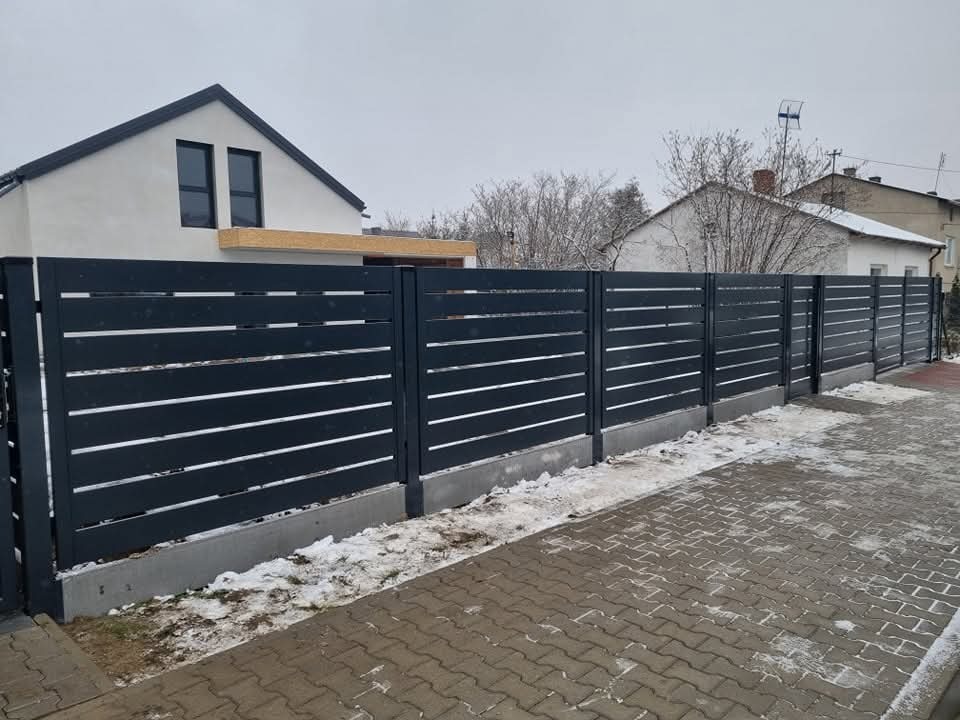 Nowoczesne, poziome, antracytowe ogrodzenie metalowe z białymi akcentami, oddzielające posesję od chodnika z kostki. Zimowy krajobraz z resztkami śniegu.