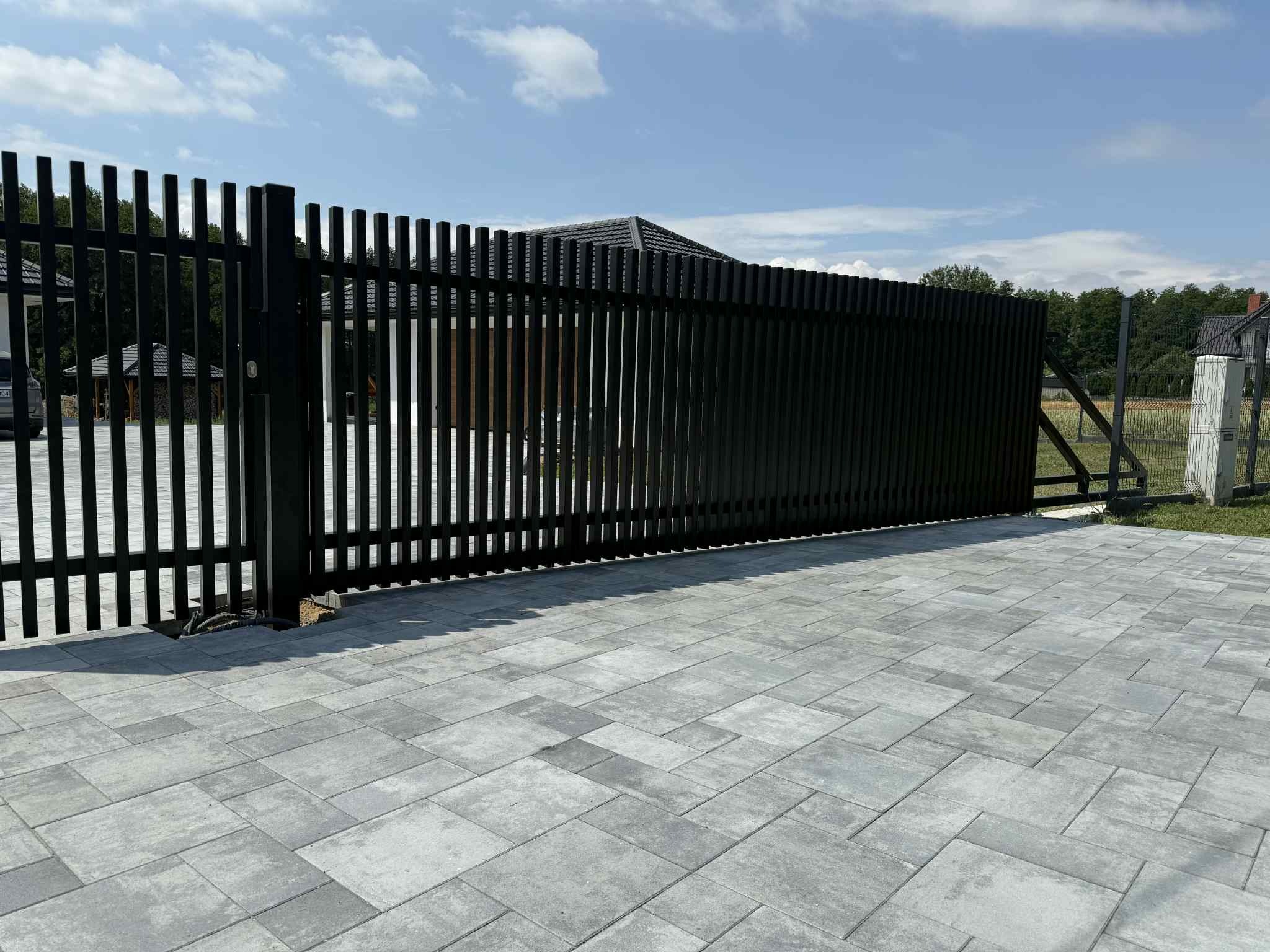 Nowoczesna, przesuwna brama wjazdowa z pionowych, czarnych profili, na tle kostki brukowej i zieleni. Widoczne elementy automatyki bramy.