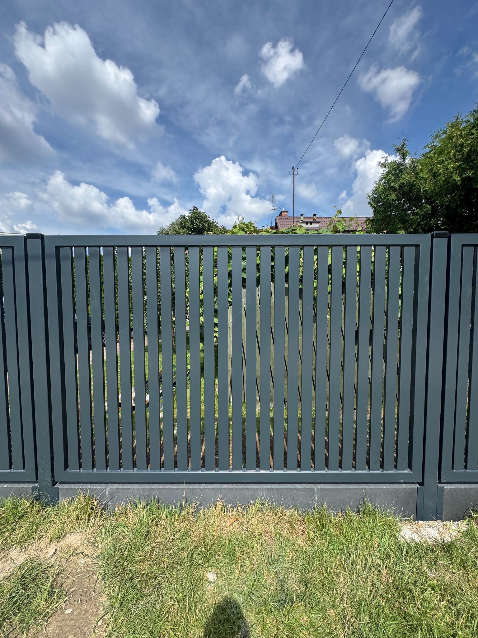 Nowoczesne, grafitowe ogrodzenie z pionowych paneli metalowych na niskiej podmurówce, z widokiem na błękitne niebo z chmurami i zieleń w tle.