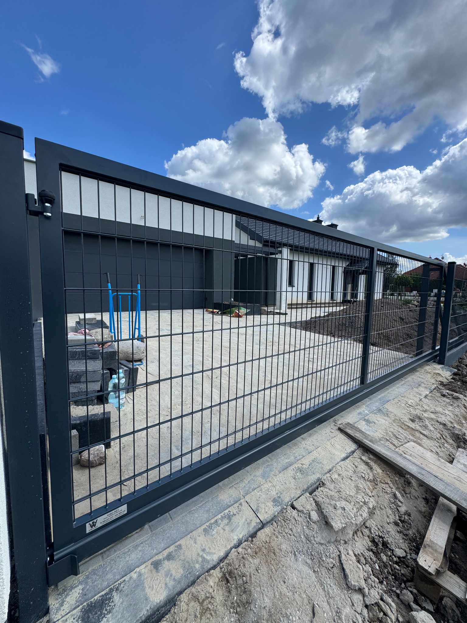 Nowoczesna, przesuwna brama wjazdowa z metalową siatką na tle domu z garażem i błękitnego nieba z chmurami. Widoczne elementy montażowe i teren w trakcie prac wykończeniowych.