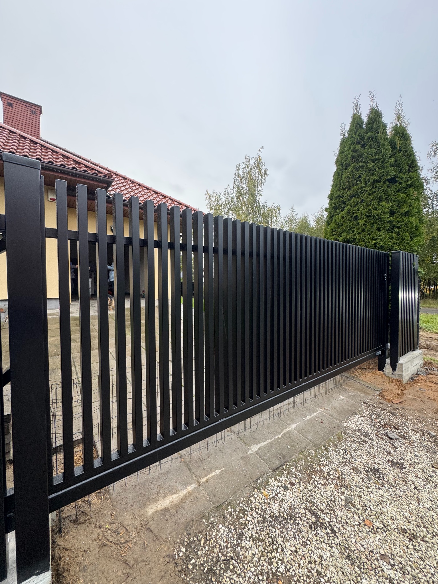 Nowoczesna, czarna brama wjazdowa z pionowych profili, zamontowana na posesji z kostką brukową i zielenią w tle. Minimalistyczny design, solidne wykonanie.