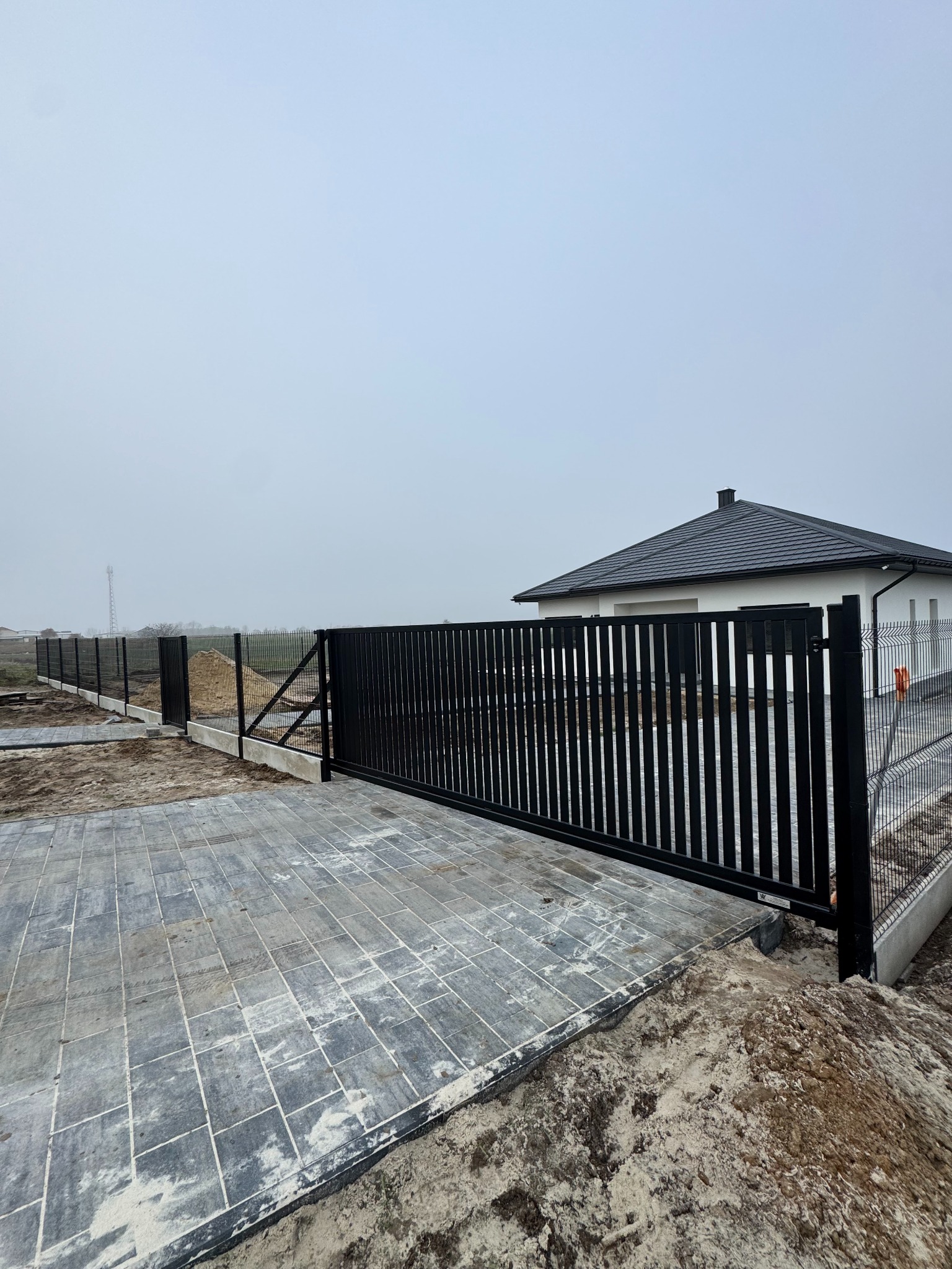 Nowoczesna, przesuwna brama wjazdowa z czarnych, pionowych profili, zintegrowana z ogrodzeniem panelowym na tle domu z ciemną dachówką w pochmurny dzień.