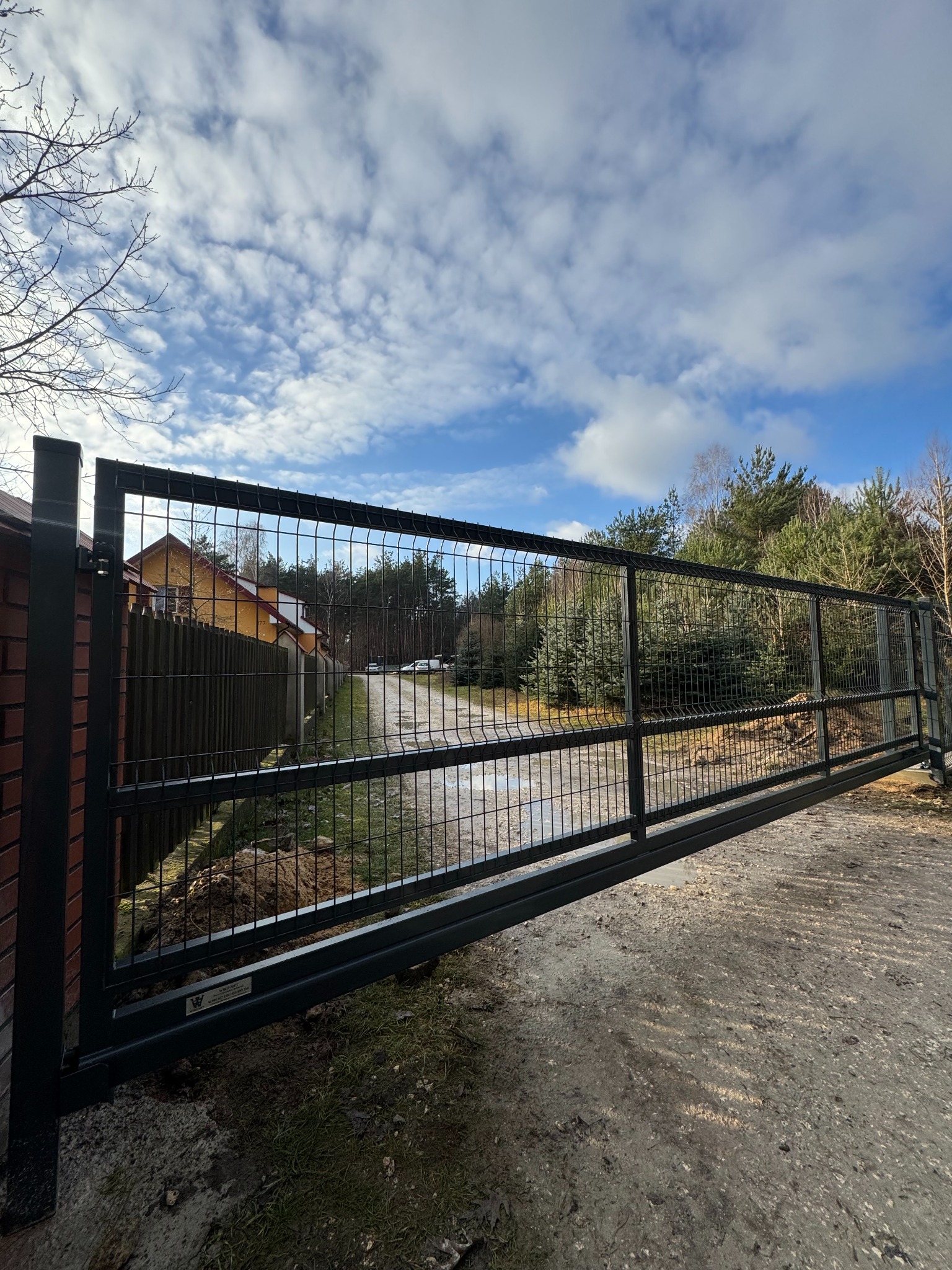 Przesuwna brama z siatki panelowej w kolorze grafitowym, widok z perspektywy gruntu, w tle dom i drzewa. Brama częściowo otwarta, widoczny mechanizm przesuwny.