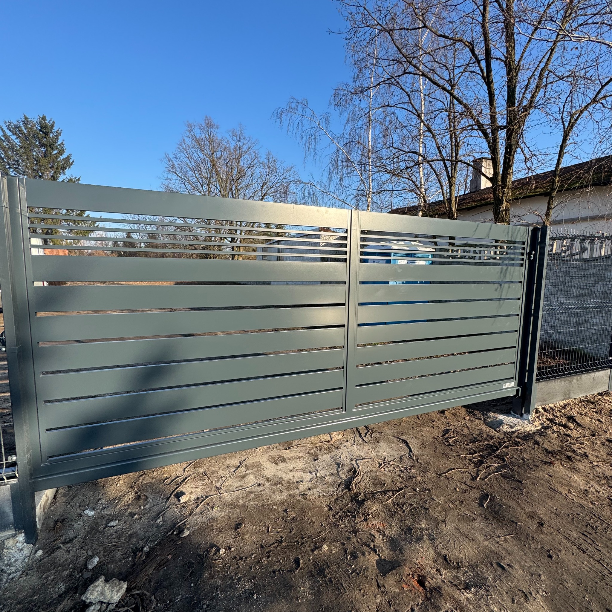 Nowoczesna, przesuwna brama wjazdowa z poziomymi panelami w kolorze grafitowym, zamontowana na posesji z widocznym fragmentem siatki ogrodzeniowej i błękitnym niebem.