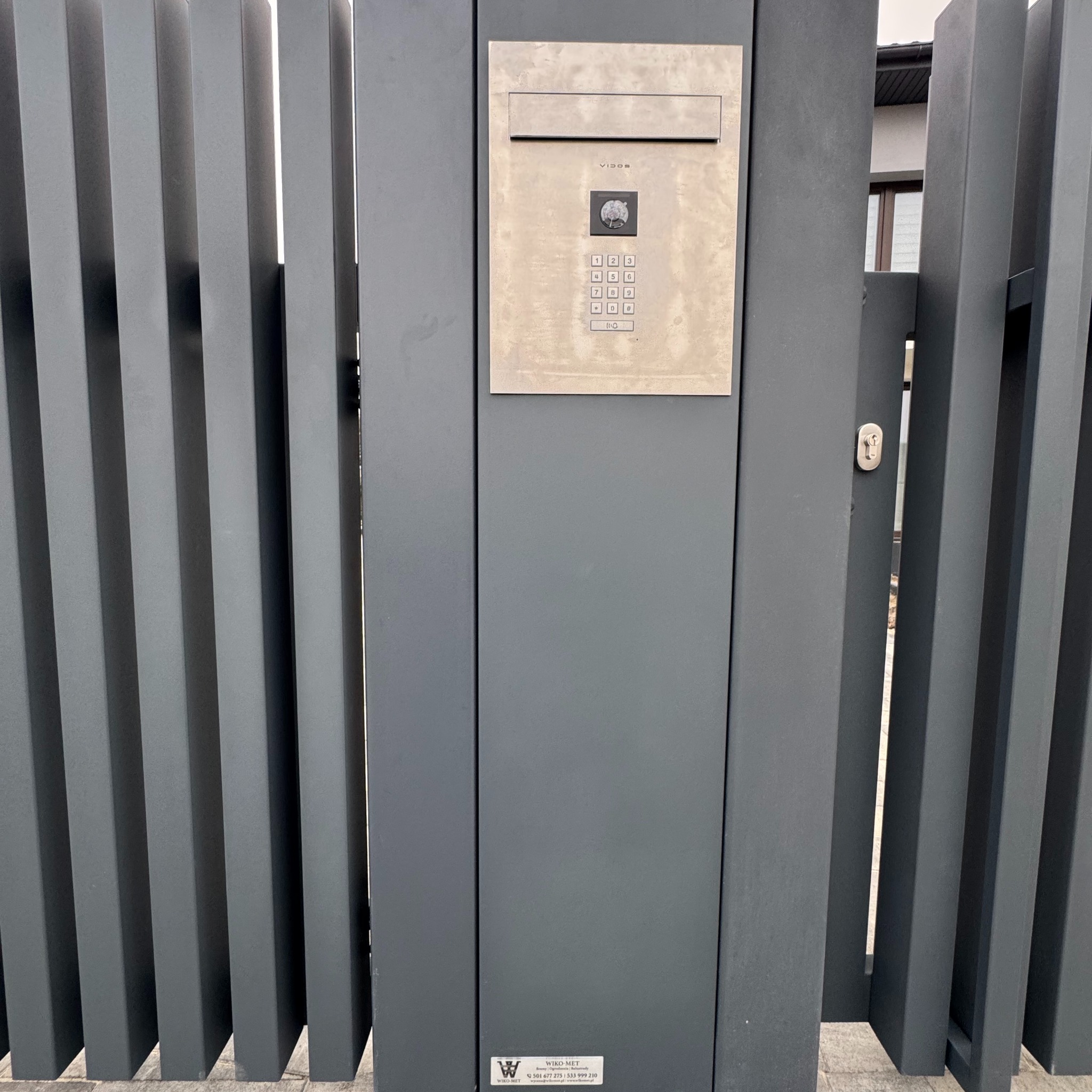 Nowoczesna, grafitowa brama wjazdowa z panelem domofonu Vidos i numerami. Minimalistyczny design, pionowe profile. Widoczny fragment domu w tle.