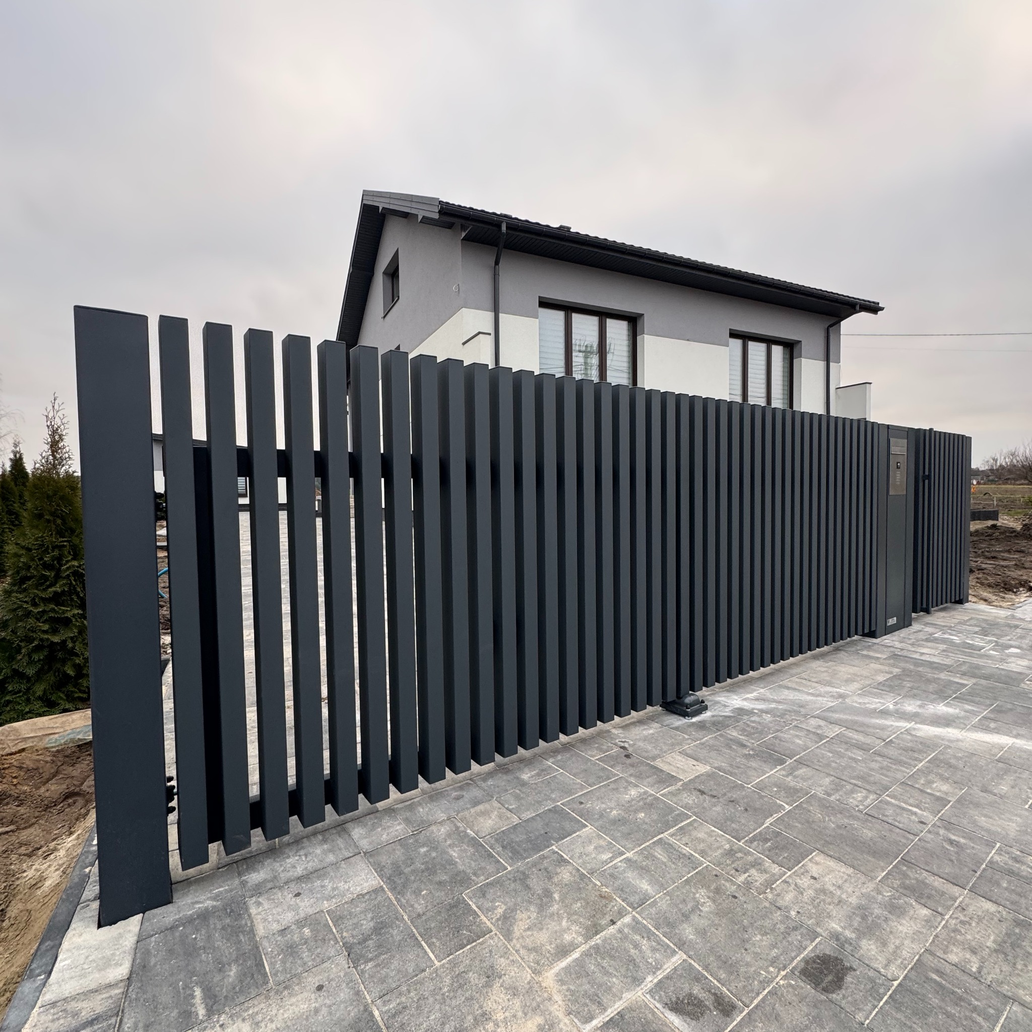 Nowoczesne, grafitowe ogrodzenie posesji z pionowych, prostokątnych profili, z furtką z domofonem, na tle domu z szarą elewacją i kostki brukowej.