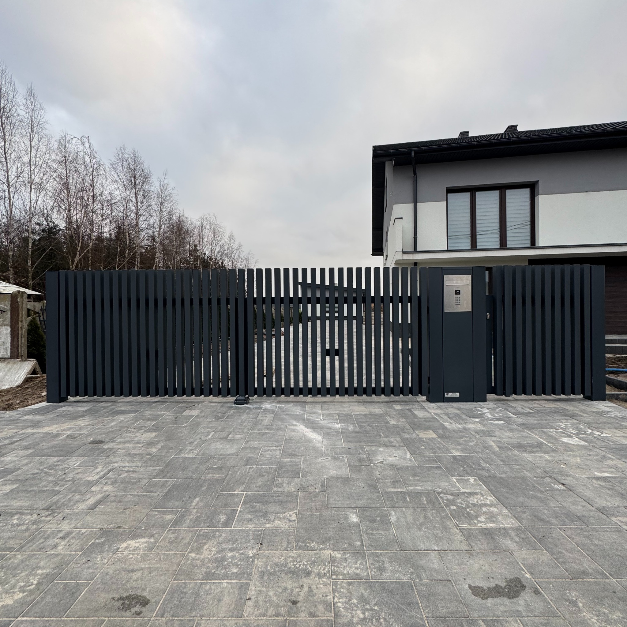 Nowoczesna, grafitowa brama wjazdowa z pionowych profili, zintegrowana ze skrzynką na listy i domofonem. Tło stanowi dom z szarą elewacją i kostka brukowa.