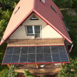 Widok z góry na dom z czerwonym dachem i panelami fotowoltaicznymi zamontowanymi na balkonie, obok powiewa flaga Polski.